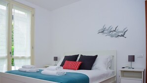 A neatly made bed with white and teal bedding, a red patterned pillow, and a white headboard. There are two bedside lamps and a wall-mounted light fixture. - Blue Bellagio by Wonderful Italy (Bellagio)