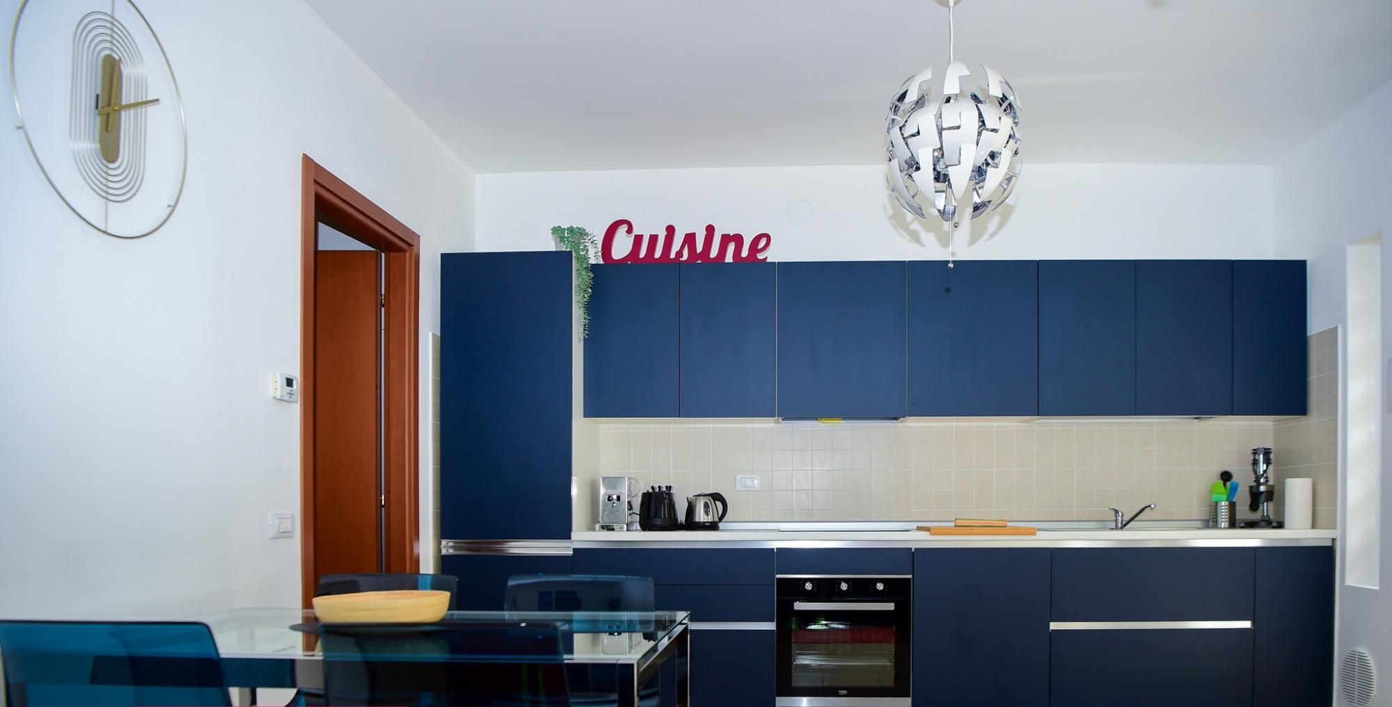 Une cuisine moderne avec des meubles bleu foncé, un plan de travail blanc et un coin repas comprenant une table et des chaises en verre.