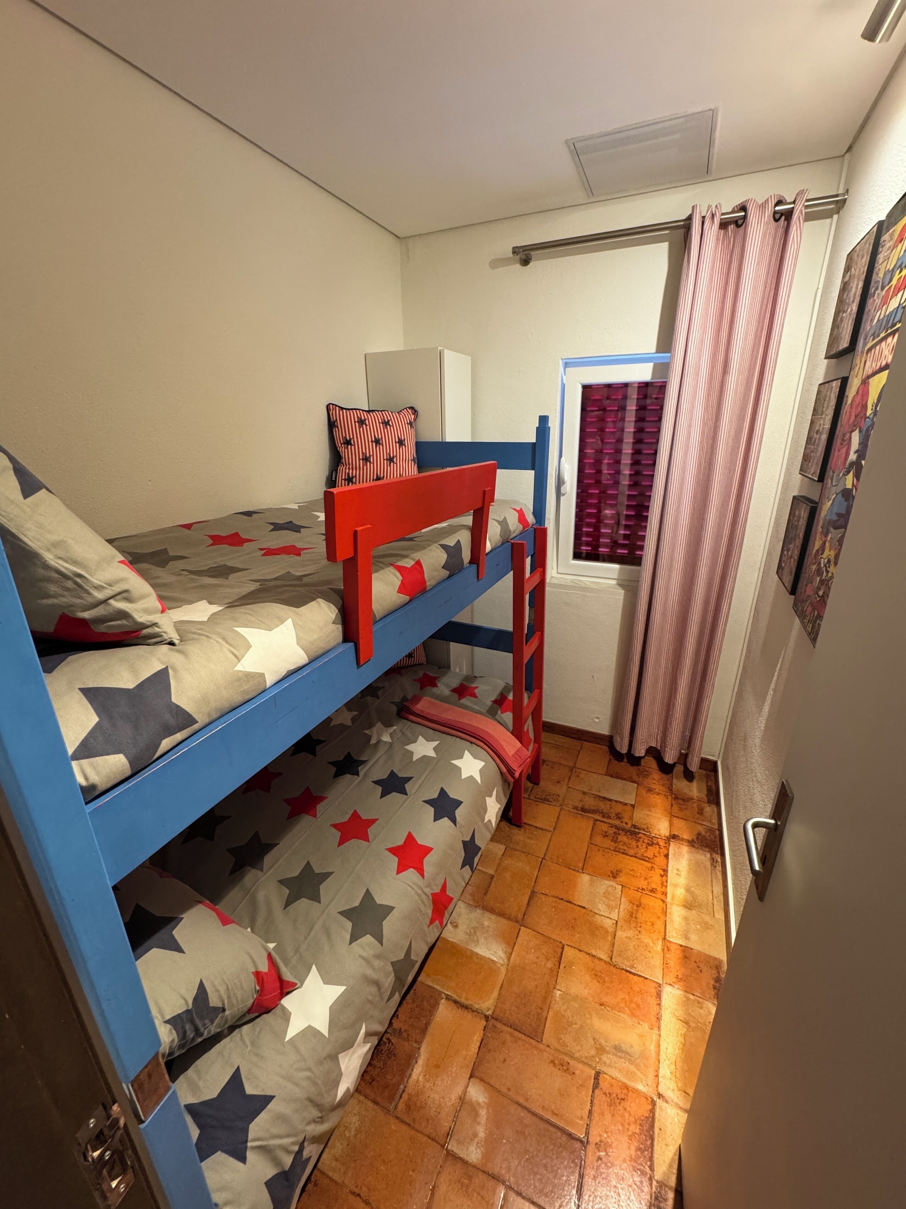 A dormitory room with bunk beds, a window with curtains, and a door with a handle.
