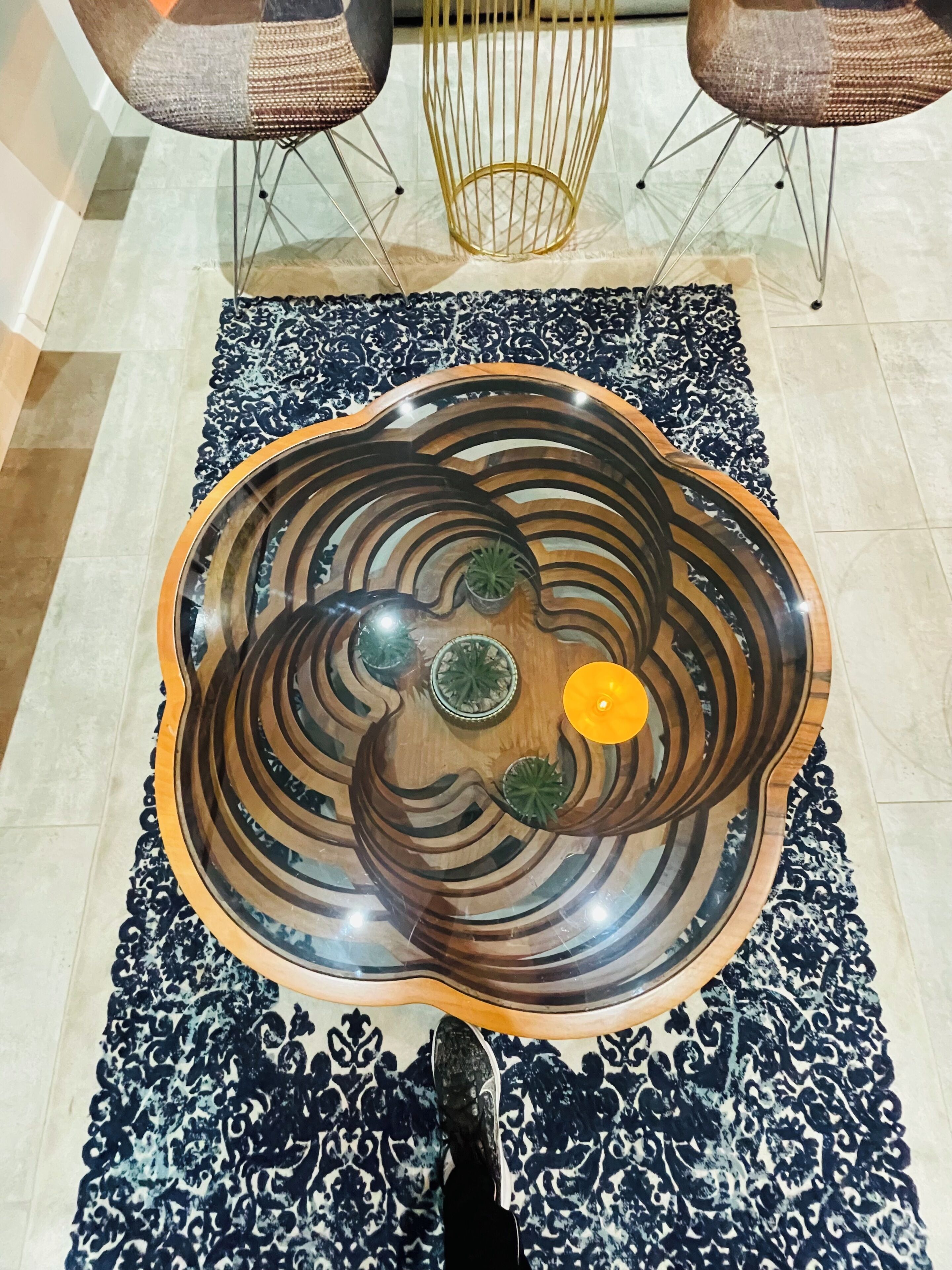 A round wooden table with a reflective surface, surrounded by a patterned rug, in a room with tiled flooring and two chairs with a woven backrest.