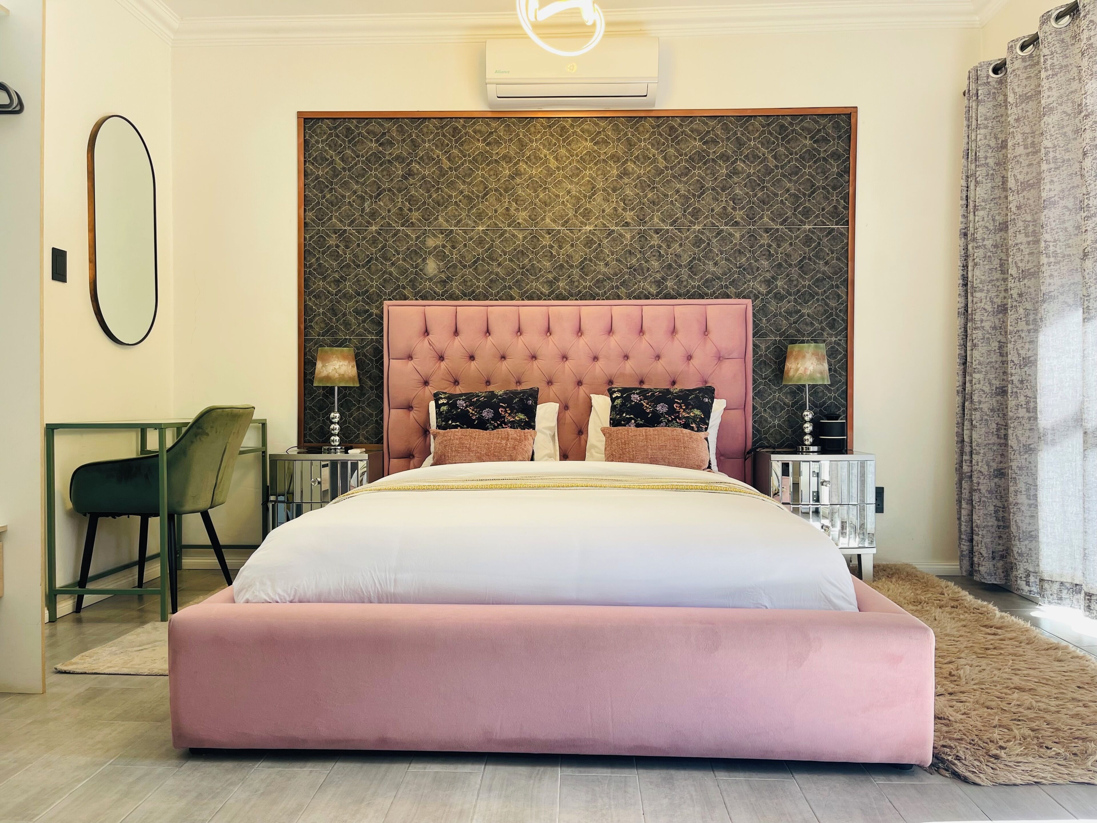 Une chambre à coucher avec une tête de lit capitonnée, un grand lit, deux tables de chevet avec des lampes, une chaise, un miroir et un mur orné d’un motif.