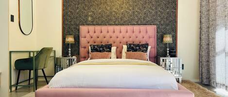 Une chambre à coucher avec une tête de lit capitonnée, un grand lit, deux tables de chevet avec des lampes, une chaise, un miroir et un mur orné d’un motif.