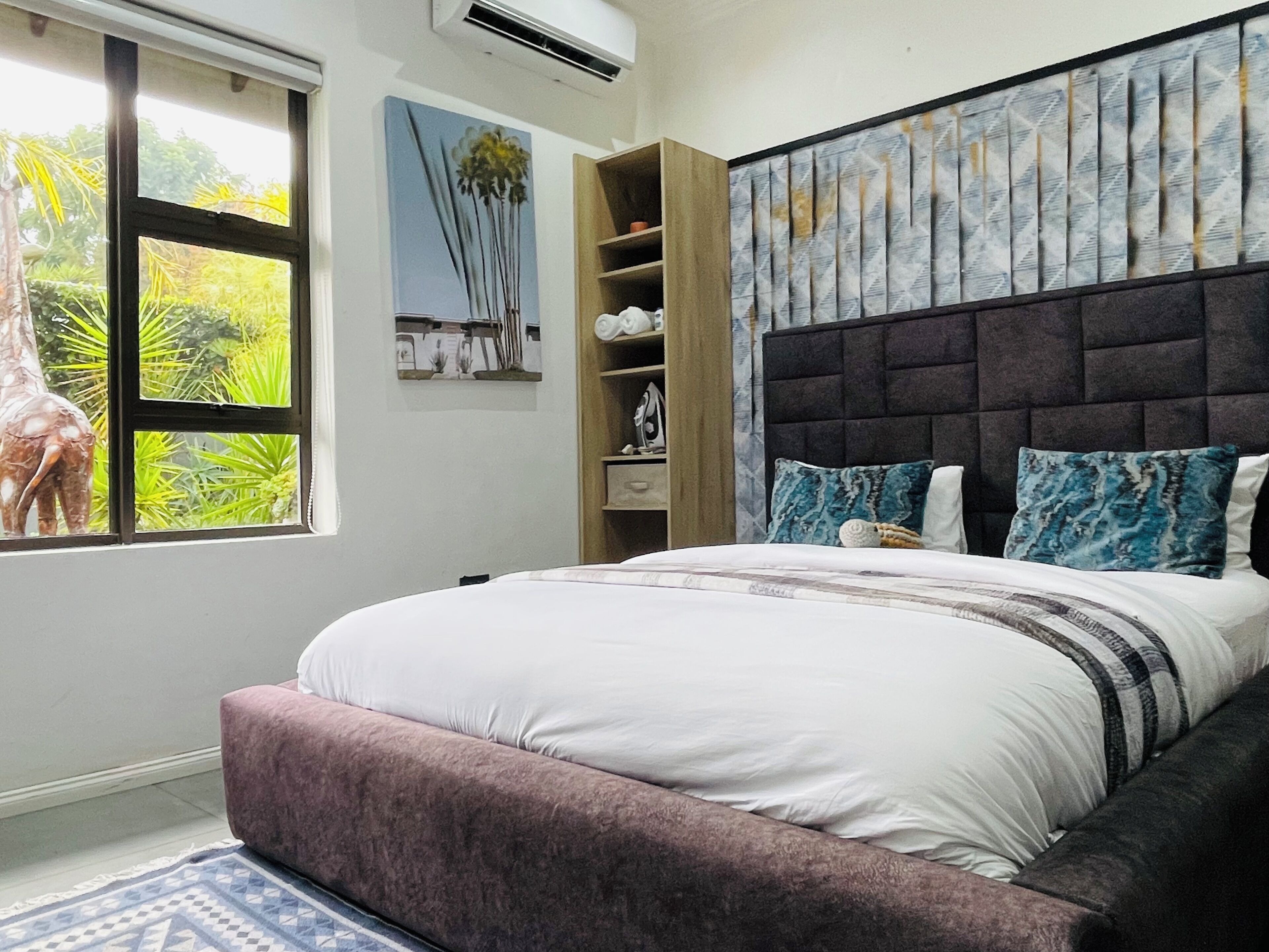 Un dormitorio con cama, ventana con vista a follaje, repisa en la pared y cabecero estampado.