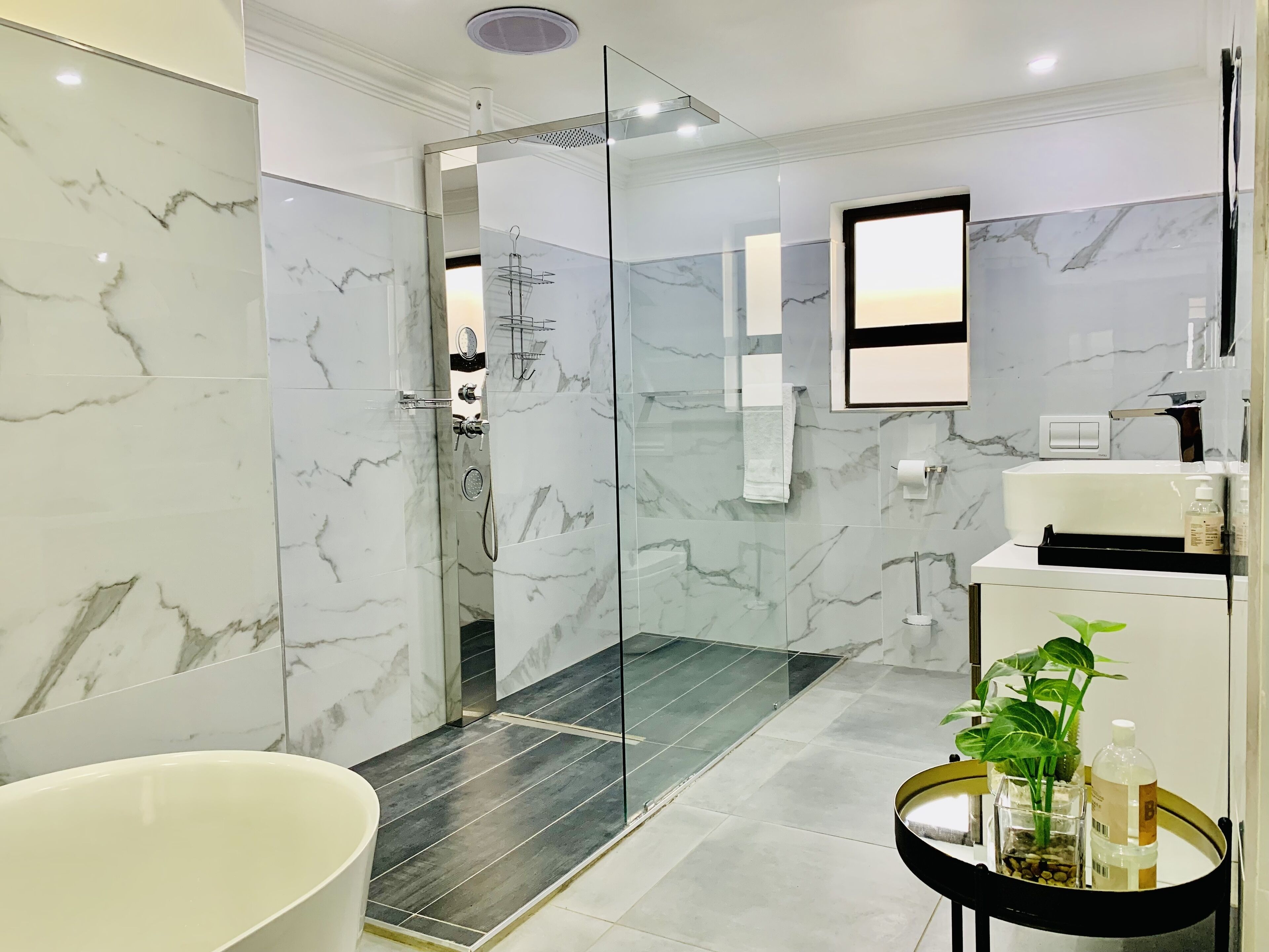 A modern bathroom with a glass-enclosed shower, marble tiles, a white sink, and a small plant on a round table.