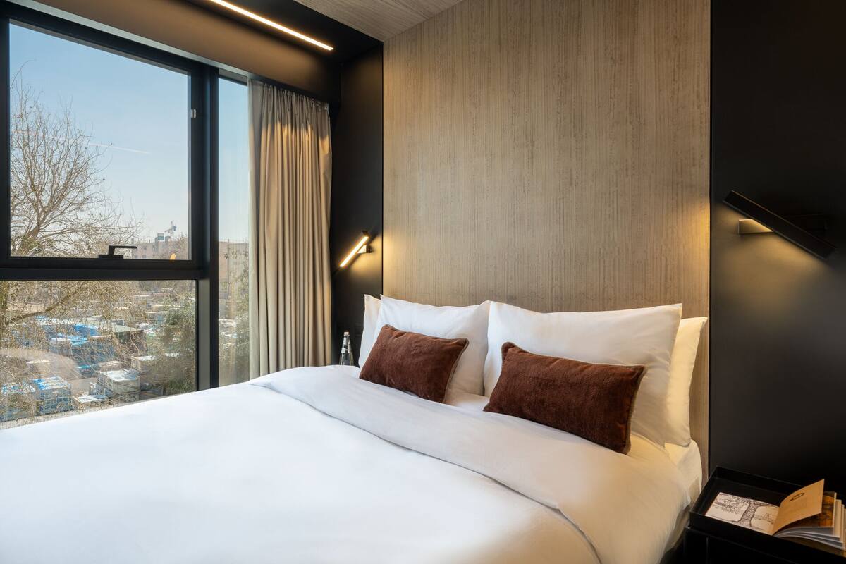 A modern hotel room with a large bed, two pillows, a nightstand, a lamp, and a window with a view of the city.