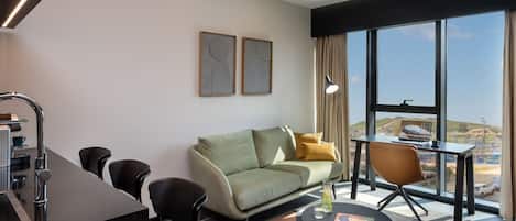 Una sala de estar moderna con un sofá, sillas, una cafetera y vistas de la ciudad a través de amplios ventanales.