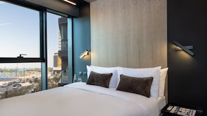 Habitación de hotel moderna con una cama grande, dos mesitas de noche y vistas a un paisaje urbano a través de amplios ventanales.