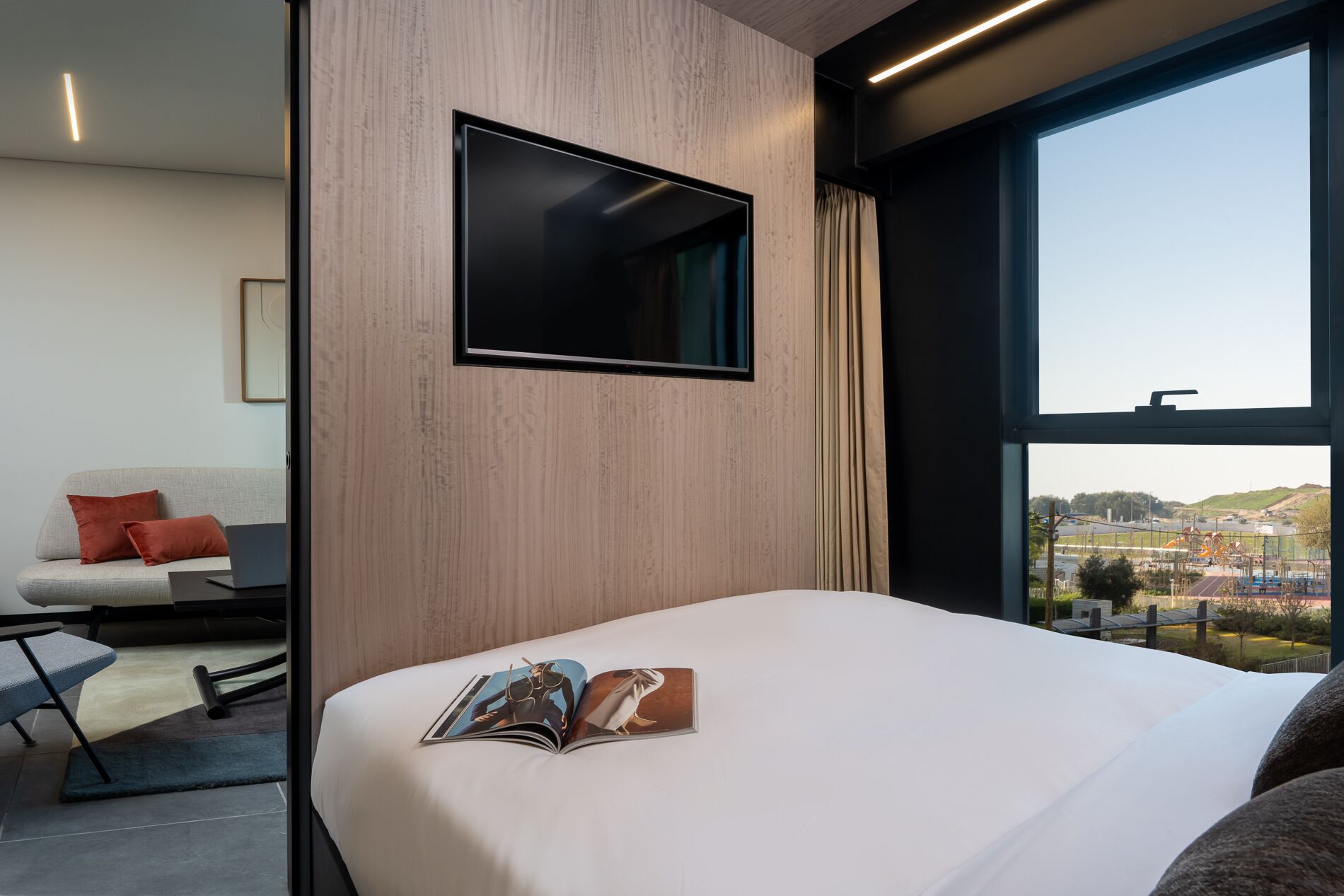 Une chambre d’hôtel moderne dotée d’un grand lit, d’un téléviseur à écran plat fixé au mur, d’un canapé et offrant une vue sur la ville grâce à de grandes fenêtres.