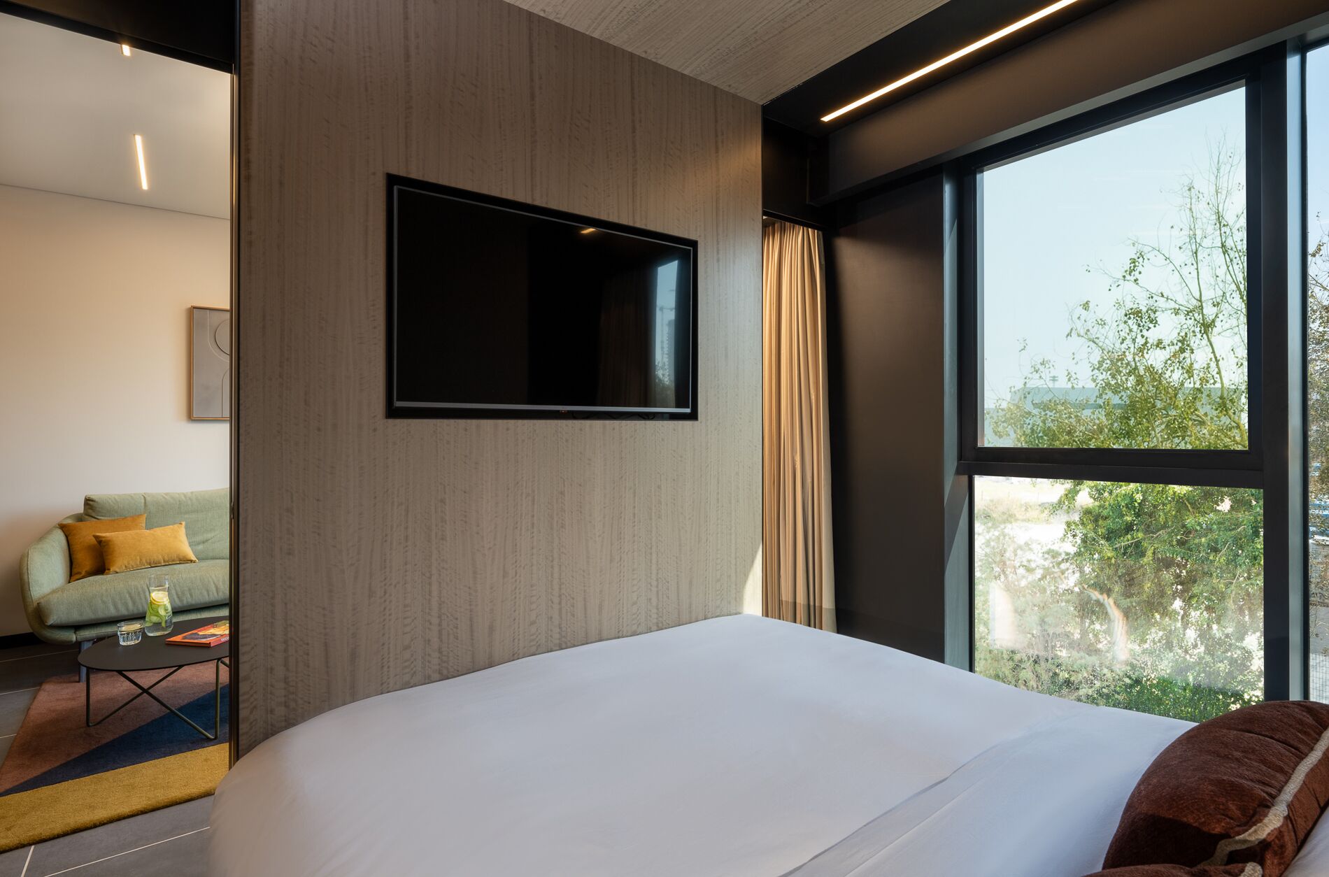Ett modernt hotellrum med en stor säng, en platt-TV som är monterad på väggen, en soffa och ett stort fönster med utsikt över träd.