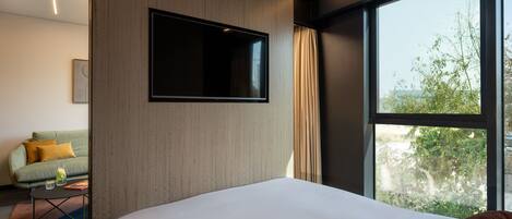 A modern hotel room with a large bed, a flat-screen TV mounted on the wall, a sofa, and a large window offering a view of trees.
