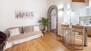 Un espacio de sala acogedor con piso de madera, un sofá, una mesa de comedor con sillas y una zona de cocina con un arco de piedra.