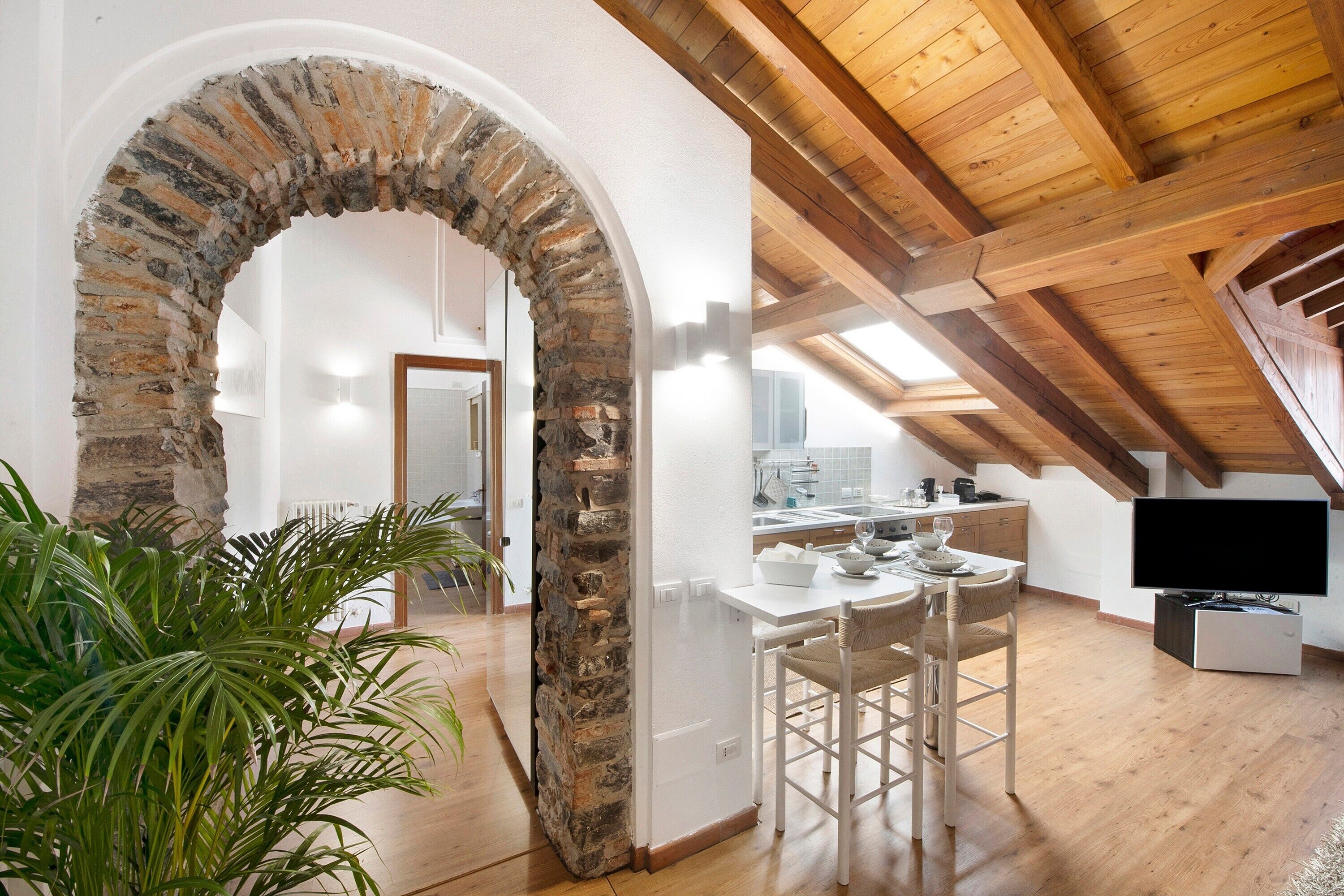 Una stanza in stile loft con un arco in pietra, travi in legno, zona pranzo con TV e cucina sullo sfondo.