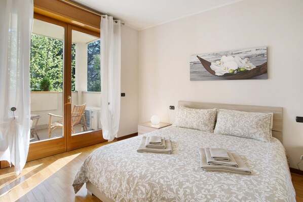 A bedroom with a bed, a nightstand, a lamp, a window with curtains, and a painting on the wall.