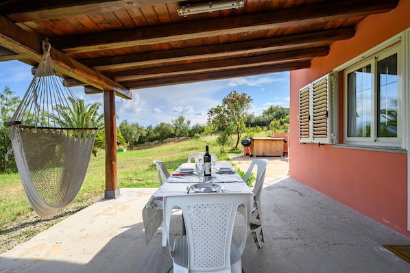 An outdoor dining area with a table set for two, surrounded by white chairs, under a wooden pergola.