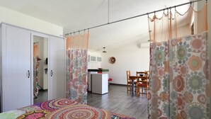 Ein Schlafzimmer mit Bett, Vorhängen, einem Kleiderschrank, einem Essbereich mit Stühlen und einer Küche im Hintergrund.