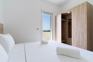 Un dormitorio con cama, ropa de cama blanca, un armario de madera y vistas a través de una puerta abierta.