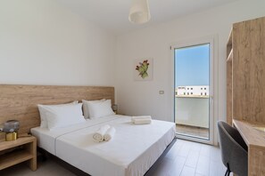 Hotellihuone, jossa on puinen sängynpääty, valkoisilla lakanoiden peitetty sänky, työpöytä tuolilla ja näkymä lasiovesta.