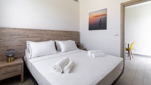 Una cama bien hecha con sábanas blancas y toallas enrolladas, un cabecero de madera, una mesita de noche con una lámpara y un cuadro de un atardecer en la pared.
