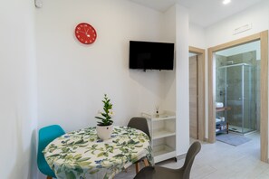 A modern room with a round table, a potted plant, a red clock, a mounted TV, and a glass shower enclosure.