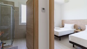 A modern hotel room with a glass shower enclosure, wooden flooring, two beds with white linens, a bedside table with a lamp, and a wooden headboard.