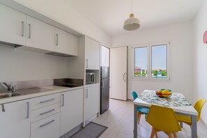 Una cocina moderna con armarios blancos, un lavamanos de acero inoxidable y un refrigerador. Hay una mesa de comedor con sillas amarillas y un frutero.