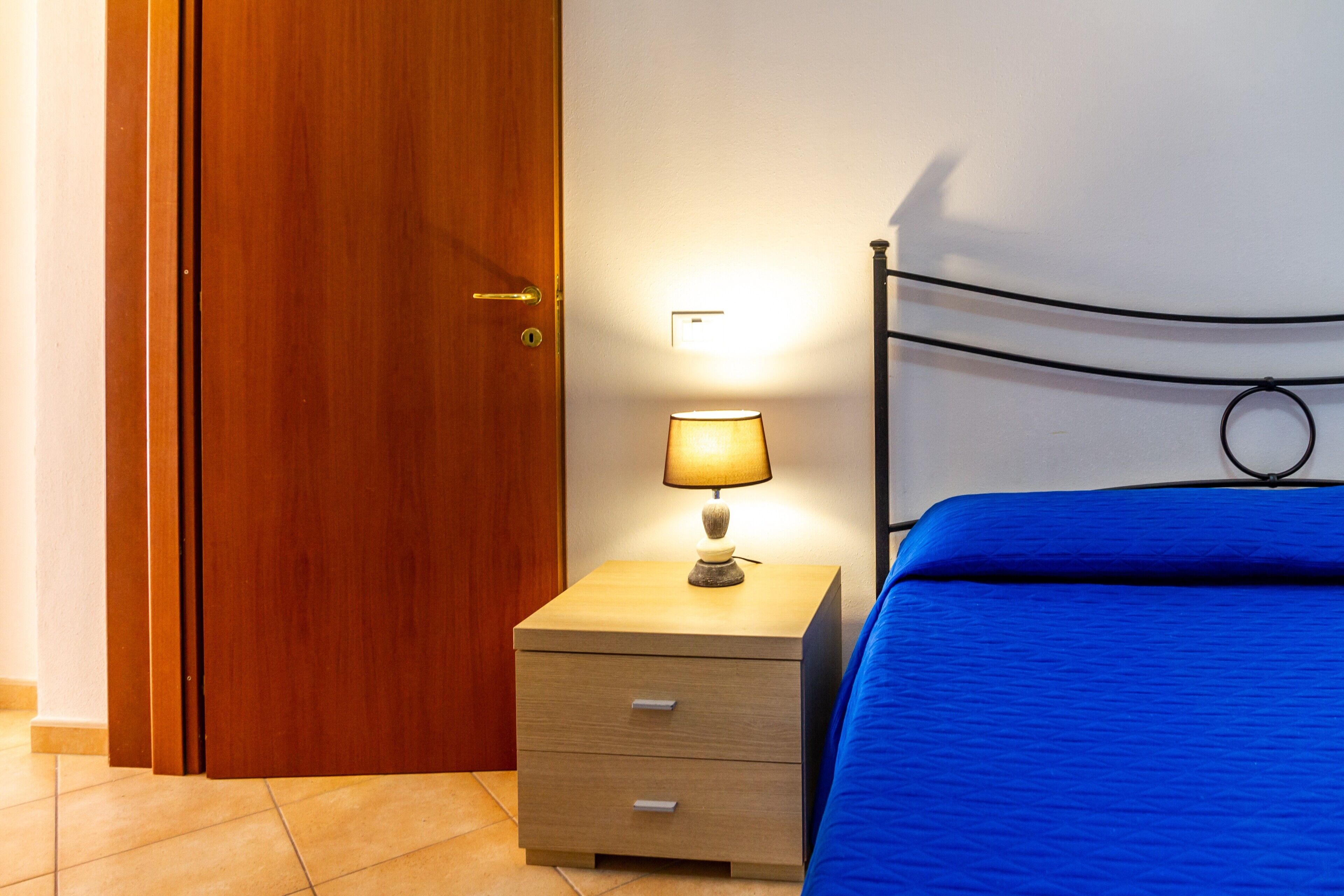 Een slaapkamer met een houten deur, een nachtkastje met een lamp en een blauw bed.