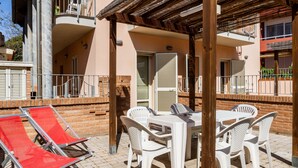 Um pátio externo com mesa e cadeiras, uma poltrona reclinável vermelha e uma pérgola de madeira.