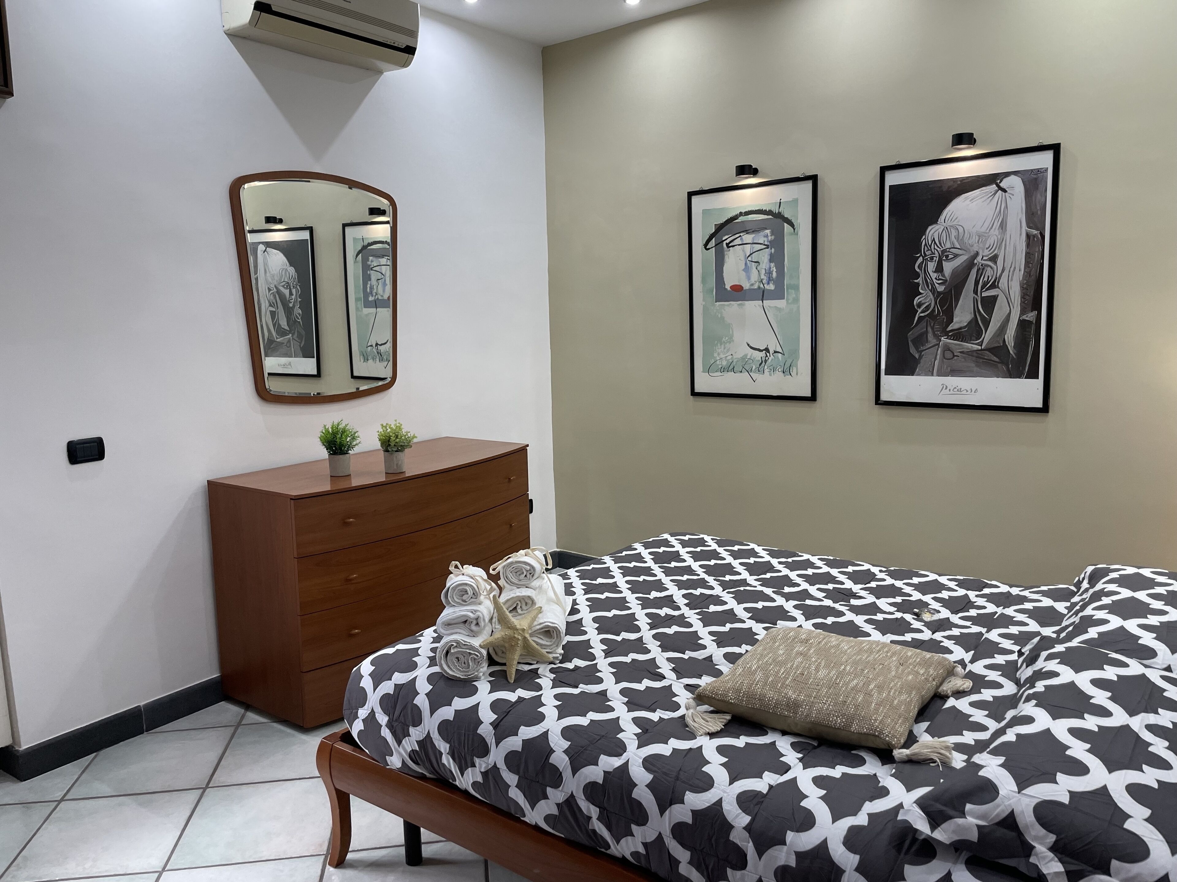 A bedroom with a bed, a dresser, a mirror, and two framed artworks on the wall.