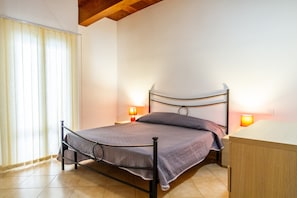 Una cama individual con estructura metálica, dos lámparas de noche, una mesita de noche de madera y una ventana con persianas.