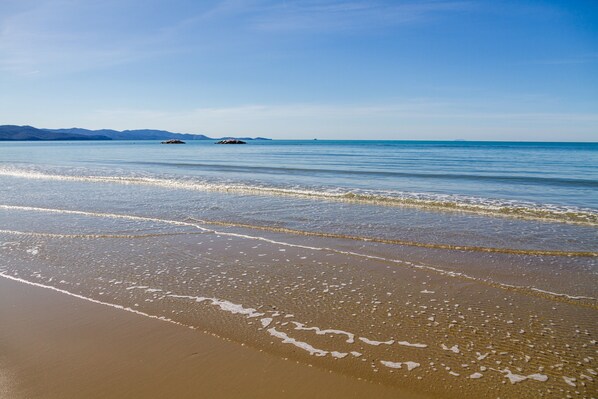 Una playa con aguas tranquilas, cielo despejado y montañas a lo lejos.