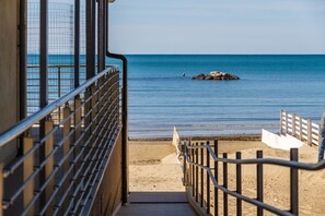 Vista de una playa desde un edificio con baranda de metal, una playa arenosa y un promontorio rocoso en el agua.