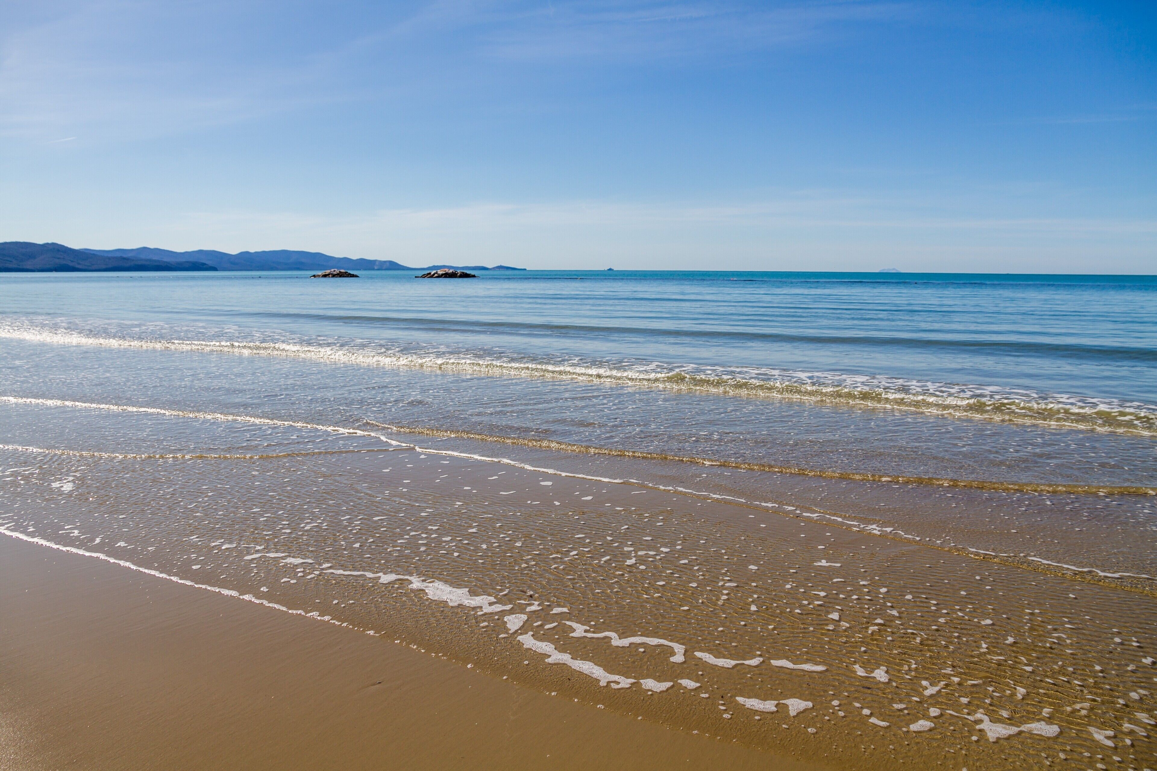 A beach with gentle waves, a clear sky, and distant mountains.