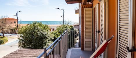 A balcony with a red chair, a view of the sea, and a building with shutters.