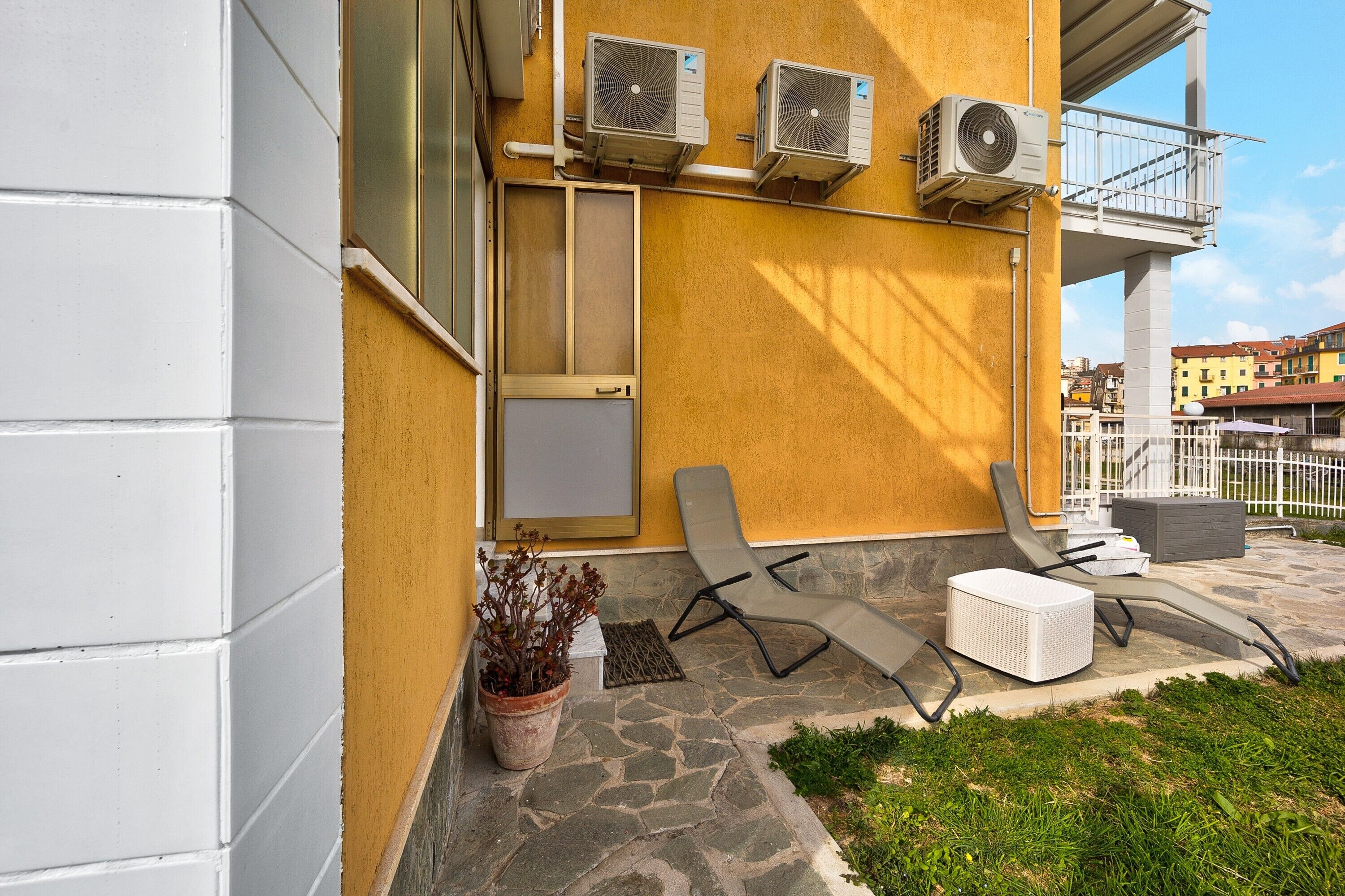 A building with air conditioning units, a balcony, a potted plant, and outdoor seating.