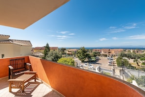 Een balkon met een houten bankje, uitzicht op een woonwijk en een heldere lucht.