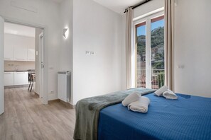 A bedroom with a bed, towels, a radiator, a window with curtains, and a view of buildings.