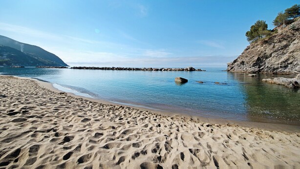 A beach with clear water, a rocky shoreline, and a mountain in the background.