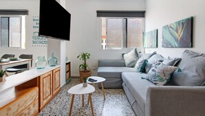 En moderne stue med en grå sofa, et fladskærms-tv, et spiseområde med et træbord og stole, samt et køkken med skabe og en bordplade.
