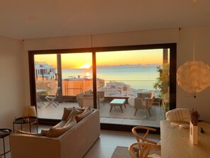 A modern living room with a large sliding glass door leading to a balcony with a view of the sunset over the sea.