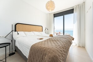 A bedroom with a bed, a bedside table, a lamp, a view of the sea, and a window with curtains.