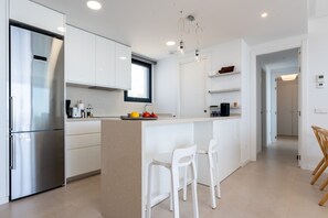 Cozinha moderna com armários brancos, geladeira de aço inoxidável, ilha central com banquetas e uma janela que oferece iluminação natural.