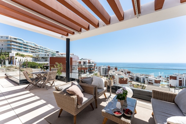 A balcony with wicker furniture, a table with a bowl of fruit, and a view of the sea and buildings.