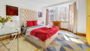 A modern hotel room with a large bed, a glass desk, a window with curtains, and a red rose painting. - Spacious Knightsbridge Apartment Near Harrods (London)