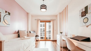 Quarto com cama, escrivaninha e cadeira. Há uma janela com cortinas, uma porta que dá para uma varanda e um lustre.