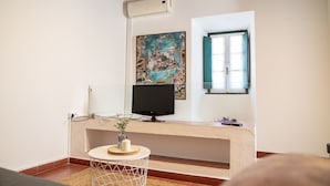 Ein Zimmer mit Fernseher auf einem Schrank, einem Bild an der Wand, einem Fenster mit blauen Fensterläden und einem kleinen runden Tisch mit einer Vase.
