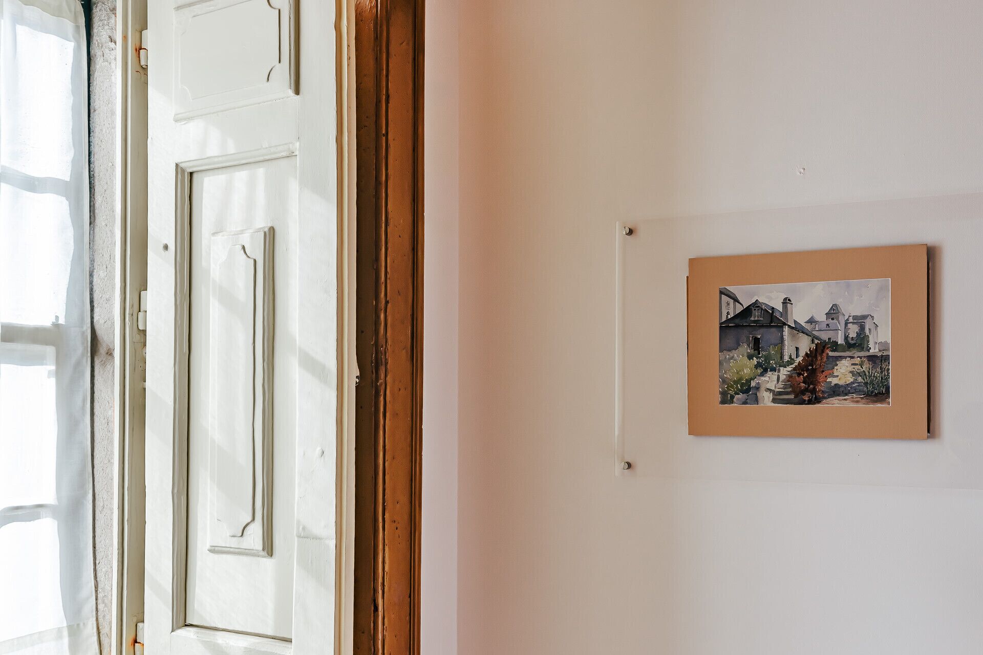 Une pièce avec une porte blanche, un tableau encadré représentant un paysage de village et une fenêtre donnant sur un immeuble.
