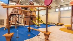 An indoor play area with a wooden climbing structure, a slide, and a swing set.