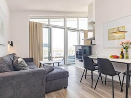 En moderne stue med en grå sofa, et spisebord med stole, et fladskærms-tv og et stort vindue med gardiner.