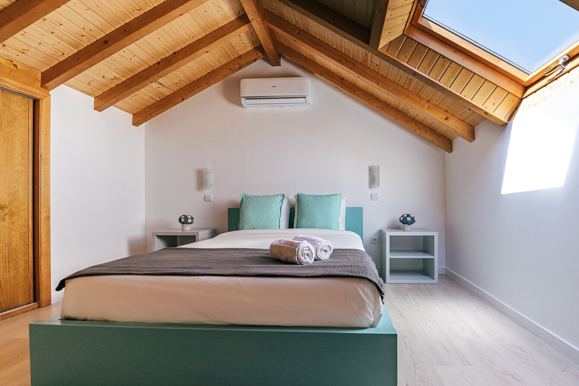 Une chambre avec un plafond en pente, un lit, deux tables de chevet, un climatiseur fixé au mur et un puits de lumière.