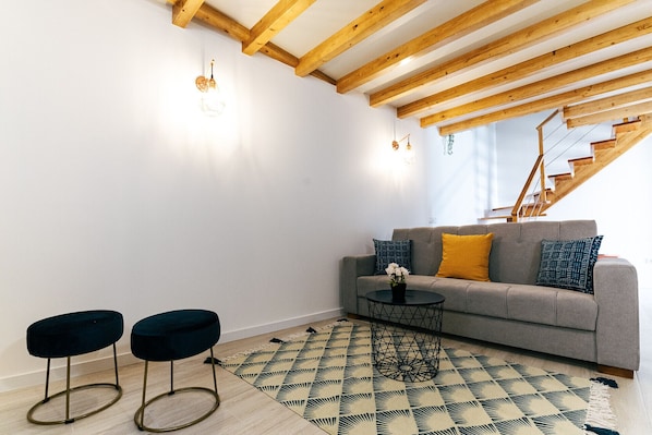 A living room with a grey sofa, two dark stools, a patterned rug, and exposed wooden beams.